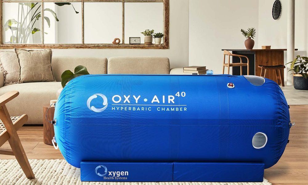 A soft-shell, tubular hyperbaric oxygen chamber in an open concept living room. The home has a sofa, chair, and dining table.